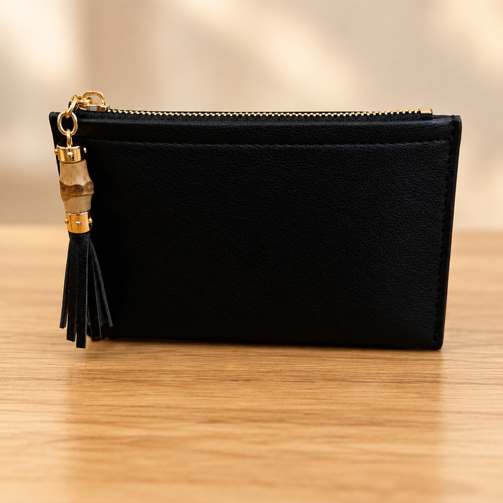 Black zippered pouch with gold hardware and tassel on a warm wooden table against a soft neutral background