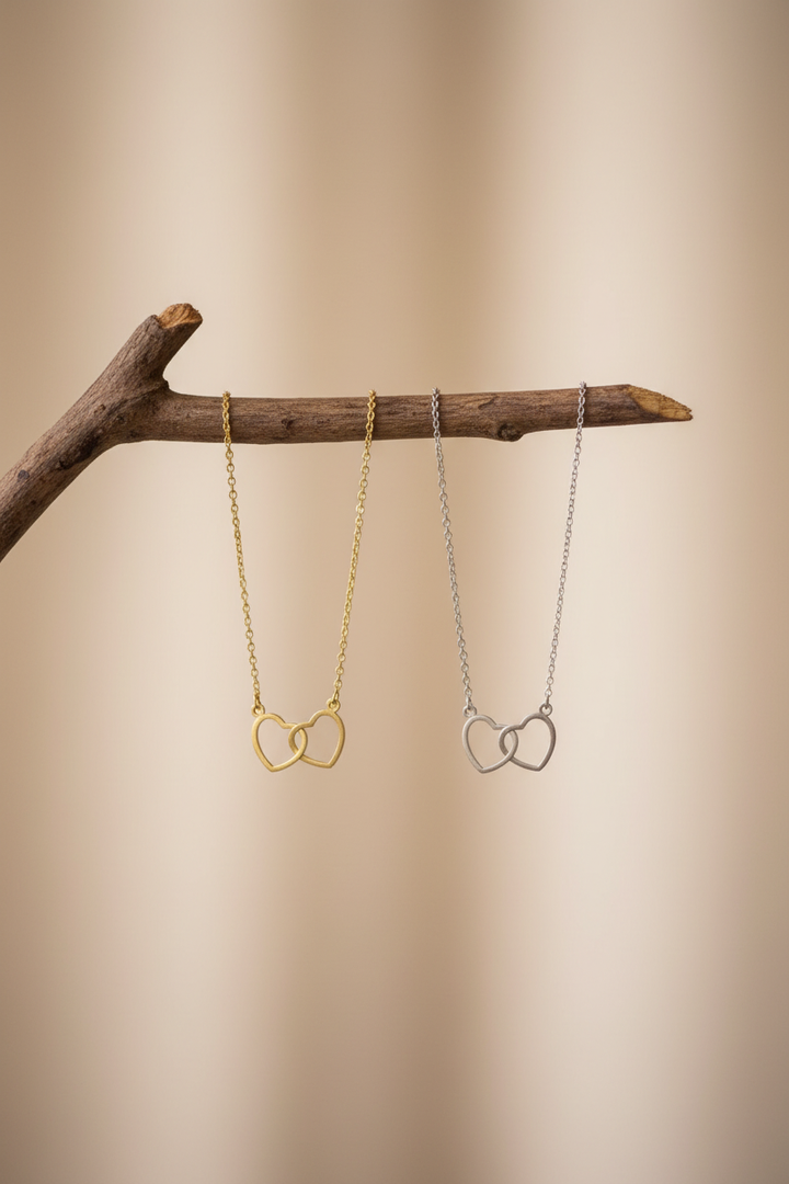 Gold and silver interlocking double heart necklaces hanging from a rustic wooden prop against a soft neutral background