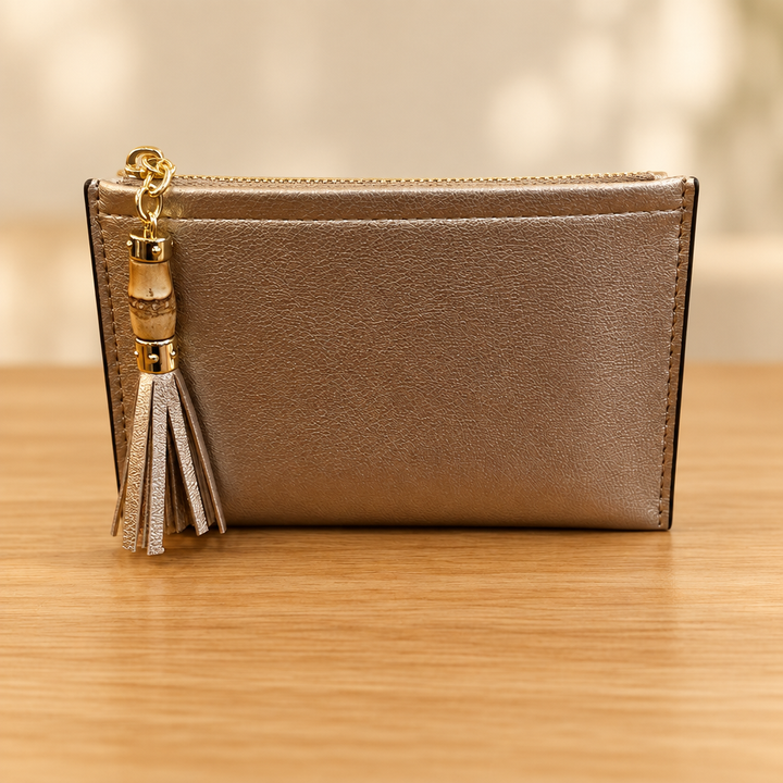 Metallic taupe zippered pouch with gold hardware and tassel on a warm wooden table against a soft neutral background