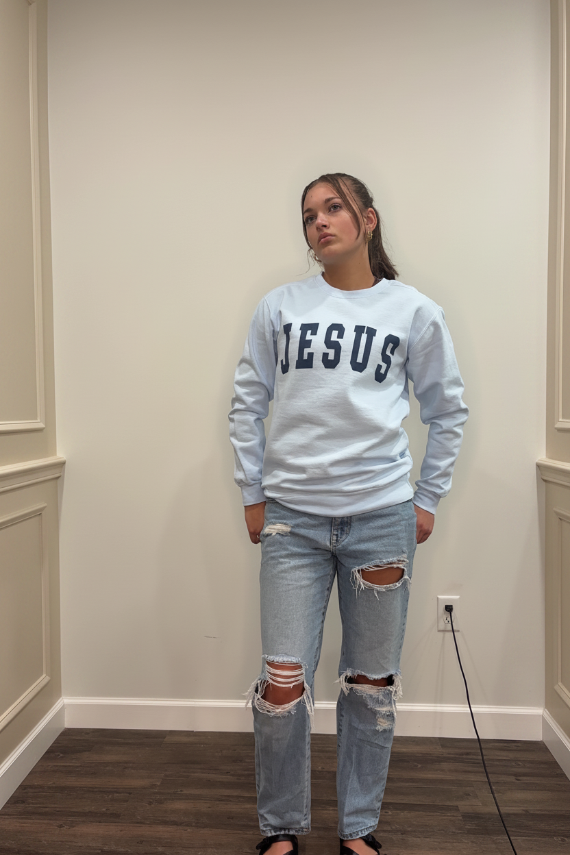Model wearing the light blue Jesus arch lettering crewneck sweatshirt in a warm neutral boutique setting, front view