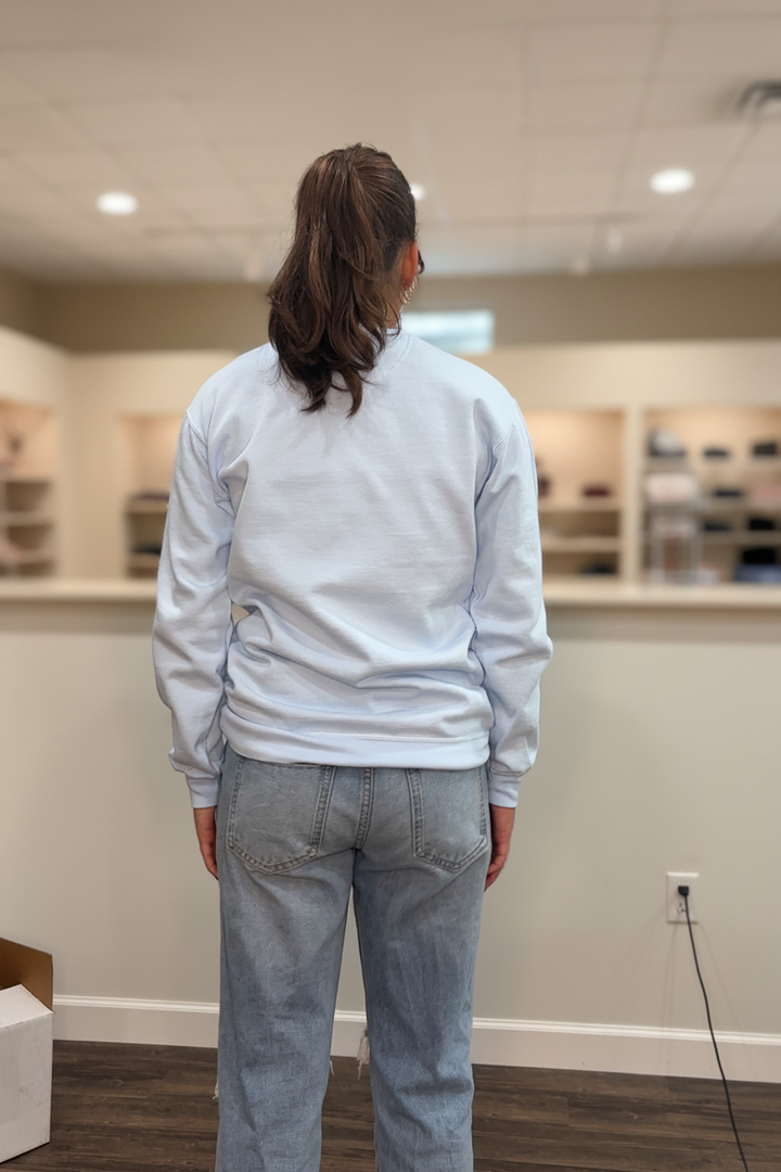 Model wearing the light blue Jesus arch lettering crewneck sweatshirt in a warm neutral boutique setting, back view with plain sweatshirt back