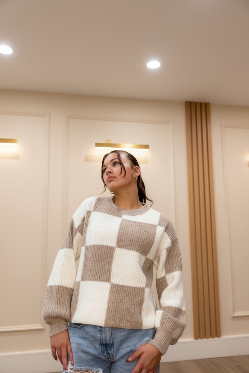 Model wearing a taupe and ivory checker crew neck sweater in a warm neutral boutique setting, front view