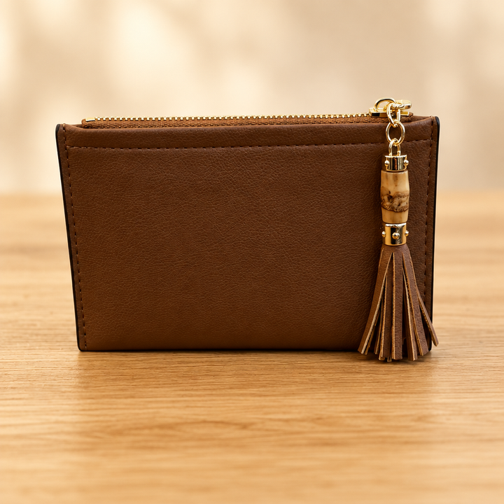 Brown zippered pouch with gold hardware and tassel on a warm wooden table against a soft neutral background